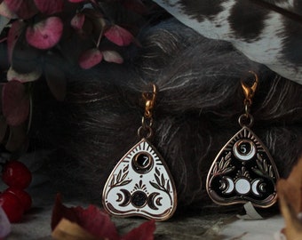 May include: Two gold-tone charm earrings with a white and black ouija board design. The charms are shaped like a triangle with a crescent moon and stars. The earrings are hanging from a feather and flowers.