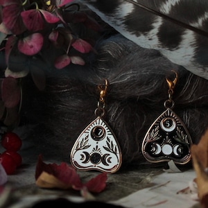 Puede incluir: Dos pendientes de aro dorados con un diseño de tablero de ouija blanco y negro. Los dijes tienen forma de triángulo con una luna creciente y estrellas. Los pendientes cuelgan de una pluma y flores.