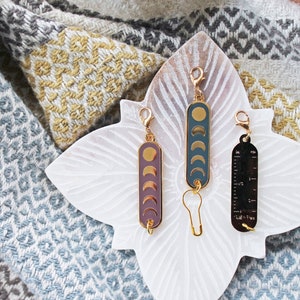 May include: Three gold-toned keychains with different designs. One keychain features a moon phase design with a dark blue background, another has a moon phase design with a purple background, and the third keychain has a ruler design with black markings.