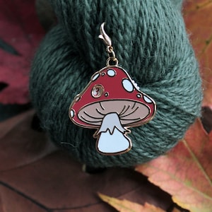 May include: A red and white mushroom shaped stitch marker with a gold metal loop. The marker is on a green ball of yarn.