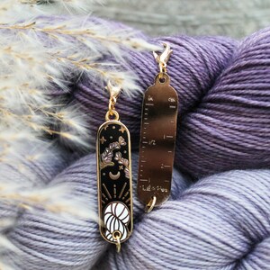 May include: Two gold-colored stitch markers with a black enamel design. One marker features a celestial design with a crescent moon, stars, and floral elements. The other has inch and centimeter markings. The markers are hanging on a skein of purple yarn.