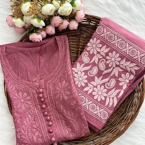 K&ouml;nnte beinhalten: Ein mauvefarbenes, besticktes Tunika und passender Stoff werden in einem Weidenkorb zusammen mit einem Strau&szlig; wei&szlig;er und rosa Blumen pr&auml;sentiert. Die Tunika hat detaillierte Blumenstickereien und eine Knopfleiste. Der Stoff hat einen floralen Rand.