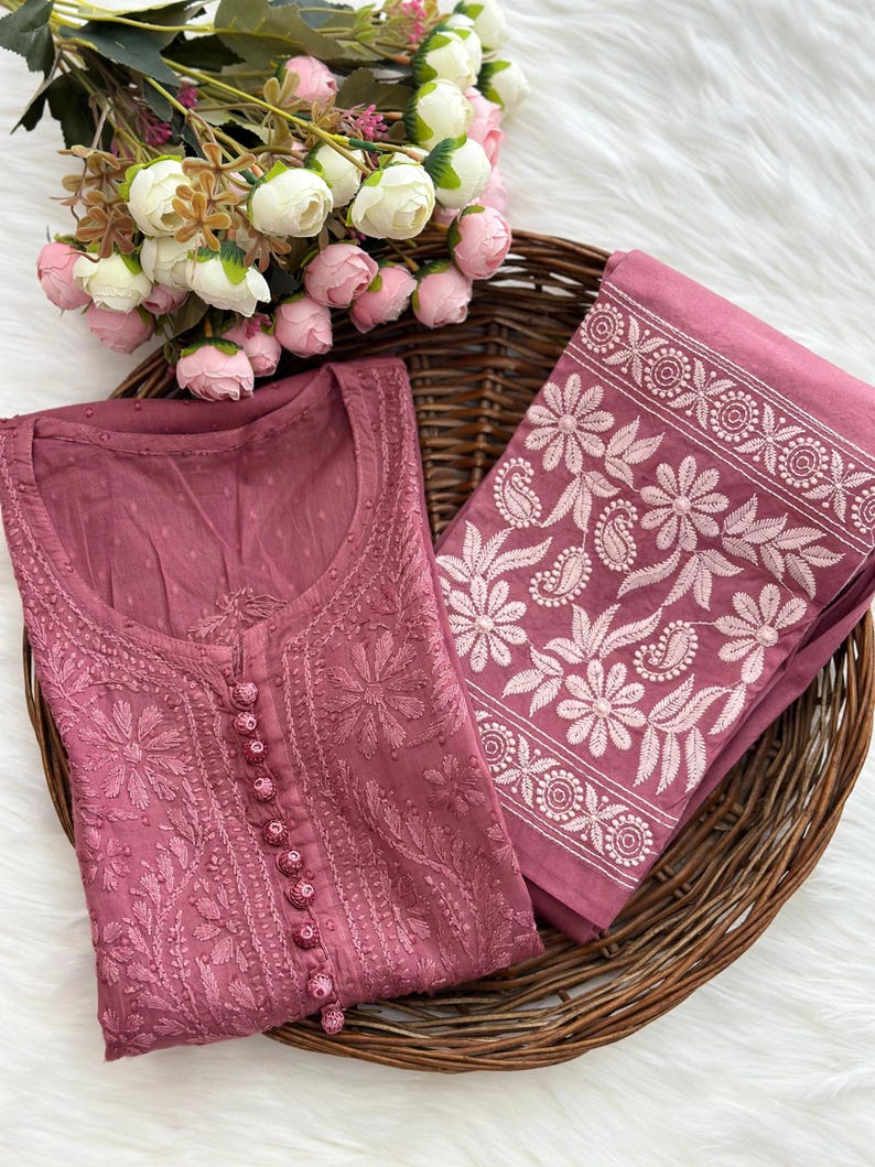 K&ouml;nnte beinhalten: Ein mauvefarbenes, besticktes Tunika und passender Stoff werden in einem Weidenkorb zusammen mit einem Strau&szlig; wei&szlig;er und rosa Blumen pr&auml;sentiert. Die Tunika hat detaillierte Blumenstickereien und eine Knopfleiste. Der Stoff hat einen floralen Rand.