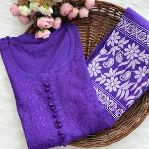 K&ouml;nnte beinhalten: Ein lila besticktes Tunika-Oberteil mit passendem Schal, pr&auml;sentiert in einem Weidenkorb. Das Oberteil hat eine Knopfleiste und detaillierte Blumenstickereien. Der Schal hat ein wei&szlig;es Blumenmuster. Rosa und wei&szlig;e Blumen sind im Hintergrund.