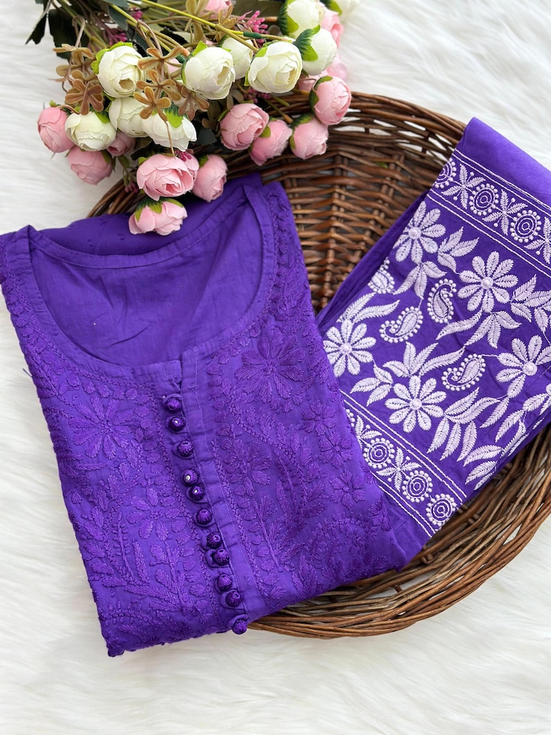 K&ouml;nnte beinhalten: Ein lila besticktes Tunika-Oberteil mit passendem Schal, pr&auml;sentiert in einem Weidenkorb. Das Oberteil hat eine Knopfleiste und detaillierte Blumenstickereien. Der Schal hat ein wei&szlig;es Blumenmuster. Rosa und wei&szlig;e Blumen sind im Hintergrund.