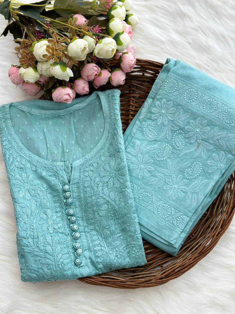 K&ouml;nnte beinhalten: Ein zweiteiliges, aquafarbenes, besticktes Outfit, bestehend aus einer Tunika und einer passenden Hose, pr&auml;sentiert in einem Weidenkorb. Die Tunika hat eine Knopfleiste und zarte Blumenstickereien. Ein Strau&szlig; rosa und wei&szlig;er Blumen befindet sich in der N&auml;he.