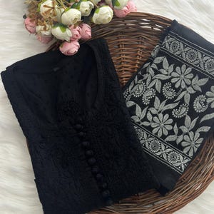 K&ouml;nnte beinhalten: Eine schwarze, bestickte Tunika mit Knopfleiste und ein passender, schwarz-wei&szlig; bestickter Schal. Die Tunika und der Schal werden auf einem Weidenkorb mit rosa und wei&szlig;en Blumen pr&auml;sentiert.