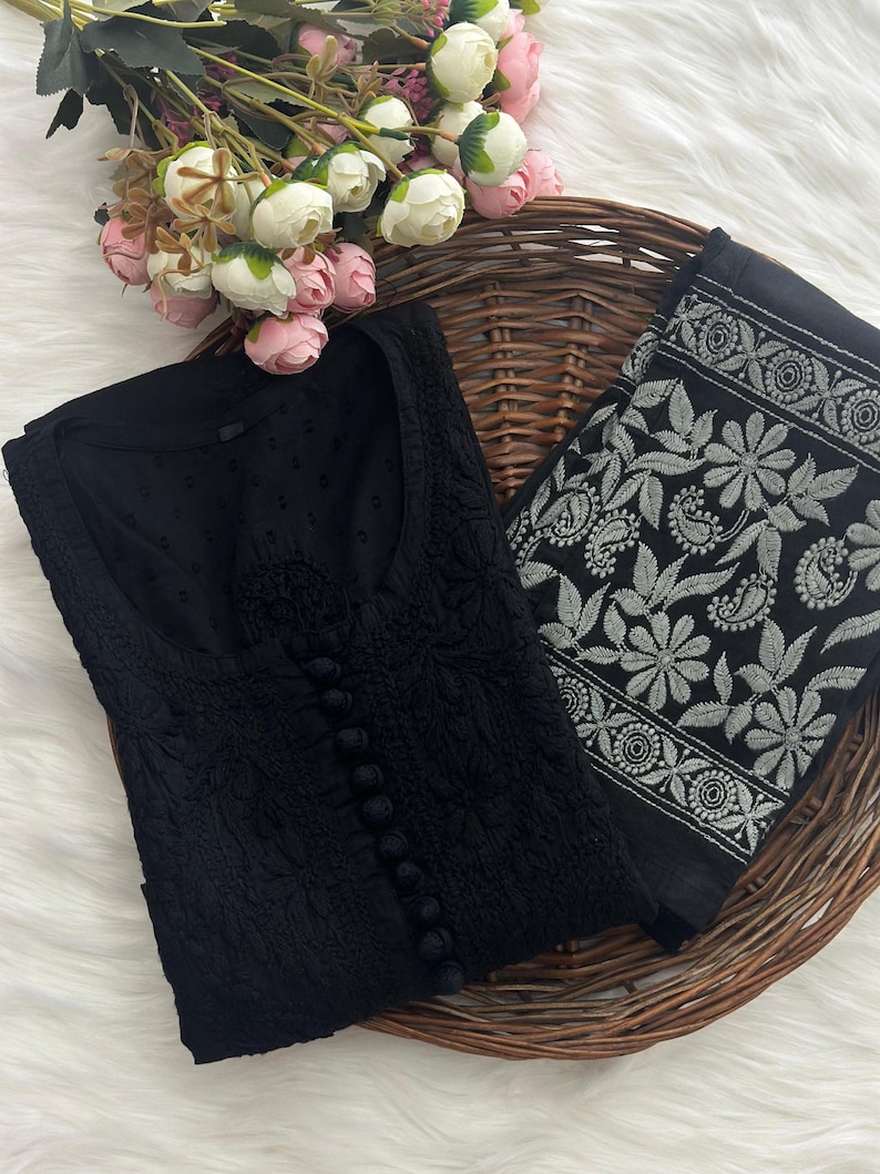 K&ouml;nnte beinhalten: Eine schwarze, bestickte Tunika mit Knopfleiste und ein passender, schwarz-wei&szlig; bestickter Schal. Die Tunika und der Schal werden auf einem Weidenkorb mit rosa und wei&szlig;en Blumen pr&auml;sentiert.