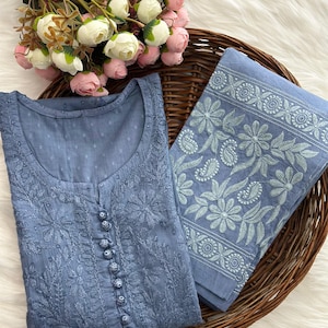 K&ouml;nnte beinhalten: Eine blaue bestickte Tunika und passender Stoff werden in einem Weidenkorb mit einem Strau&szlig; rosa und wei&szlig;er Blumen pr&auml;sentiert. Die Tunika hat eine Knopfleiste vorne und aufwendige Blumenstickereien. Der Stoff hat einen floralen Rand.