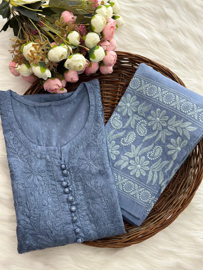 K&ouml;nnte beinhalten: Eine blaue bestickte Tunika und passender Stoff werden in einem Weidenkorb mit einem Strau&szlig; rosa und wei&szlig;er Blumen pr&auml;sentiert. Die Tunika hat eine Knopfleiste vorne und aufwendige Blumenstickereien. Der Stoff hat einen floralen Rand.