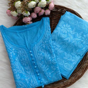 K&ouml;nnte beinhalten: Hellblaues besticktes Tunika- und Hosen-Set. Die Tunika hat einen runden Ausschnitt, eine Knopfleiste vorne und aufwendige Blumenstickereien. Die Hose hat ebenfalls passende Stickereien. Das Set wird auf einem geflochtenen Tablett mit Blumen pr&auml;sentiert.