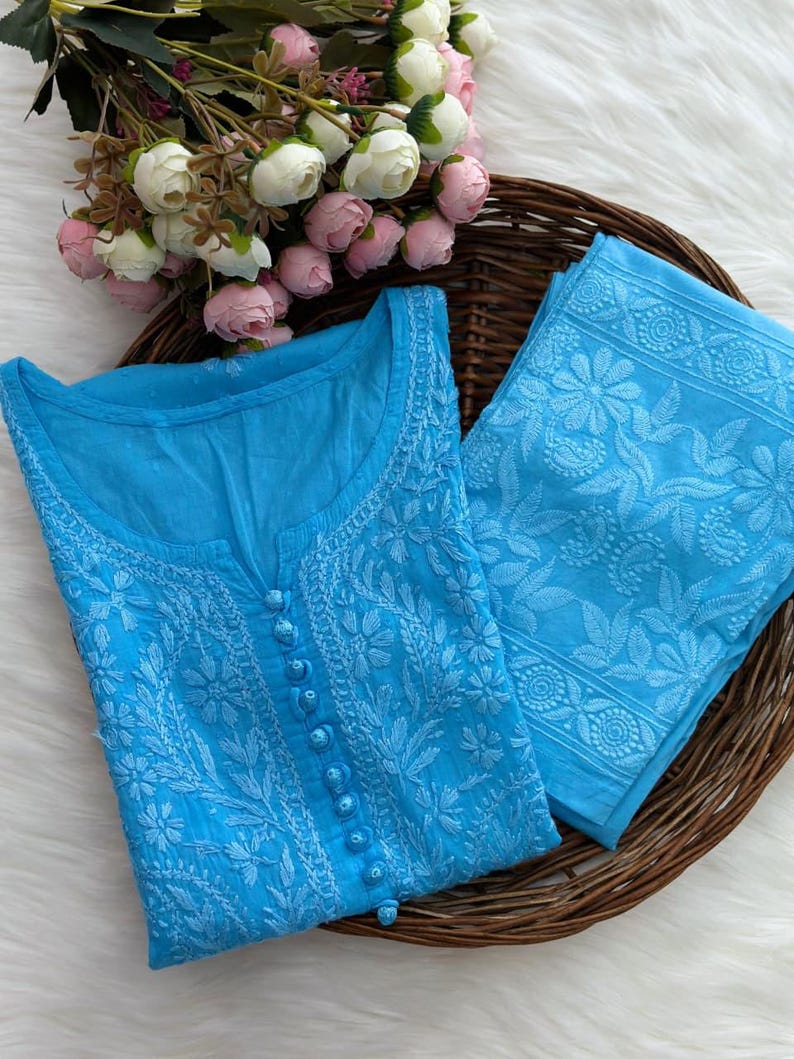 K&ouml;nnte beinhalten: Hellblaues besticktes Tunika- und Hosen-Set. Die Tunika hat einen runden Ausschnitt, eine Knopfleiste vorne und aufwendige Blumenstickereien. Die Hose hat ebenfalls passende Stickereien. Das Set wird auf einem geflochtenen Tablett mit Blumen pr&auml;sentiert.