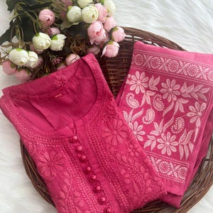 K&ouml;nnte beinhalten: Ein magentafarbenes besticktes Kurta- und Dupatta-Set. Das Kurta hat detaillierte Blumenstickereien und Knopfverschl&uuml;sse. Das Dupatta hat ein Blumenbord&uuml;rendesign. Das Set wird in einem Weidenkorb mit einem Strau&szlig; rosa und wei&szlig;er Blumen pr&auml;sentiert.