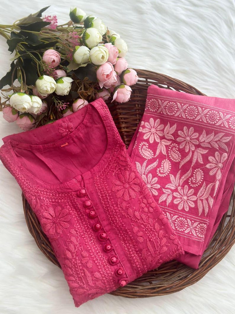 K&ouml;nnte beinhalten: Ein magentafarbenes besticktes Kurta- und Dupatta-Set. Das Kurta hat detaillierte Blumenstickereien und Knopfverschl&uuml;sse. Das Dupatta hat ein Blumenbord&uuml;rendesign. Das Set wird in einem Weidenkorb mit einem Strau&szlig; rosa und wei&szlig;er Blumen pr&auml;sentiert.