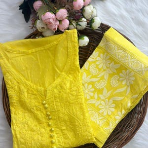 K&ouml;nnte beinhalten: Ein leuchtend gelbes besticktes Tunika- und Schal-Set. Die Tunika hat eine Knopfleiste vorne und aufwendige Blumenmuster. Der Schal zeigt ein wei&szlig;es Blumendesign. Das Set wird auf einem geflochtenen Tablett mit rosa und wei&szlig;en Blumen im Hintergrund pr&auml;sentiert.