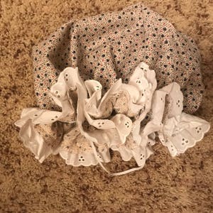 May include: A white lace bonnet with a floral print fabric lining. The bonnet has a ruffled edge and is tied with a white ribbon.