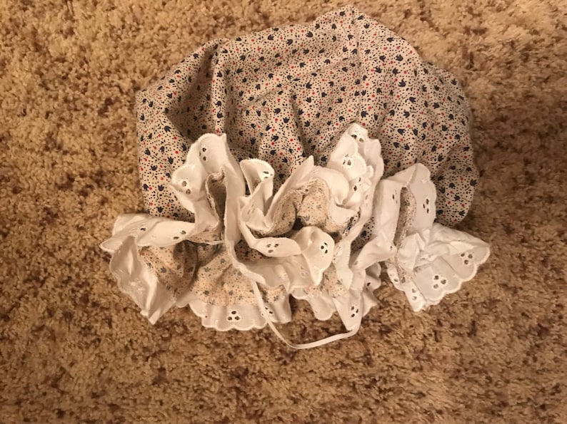 May include: A white lace bonnet with a floral print fabric lining. The bonnet has a ruffled edge and is tied with a white ribbon.