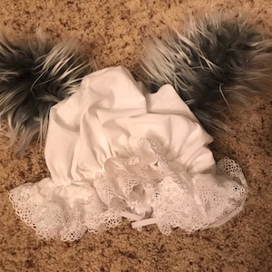 May include: A white bonnet with a lace trim and two gray and white fur tails attached.