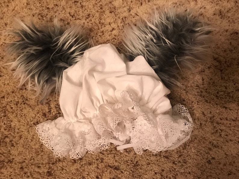 May include: A white bonnet with a lace trim and two gray and white fur tails attached.