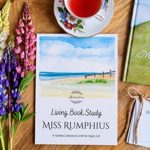 May include: A Living Book Study guide for Miss Rumphius with a watercolor beach illustration, a tea cup, a hardcover book, and a stack of sequence cards tied with twine. Three tall lupine flower stalks in white, purple, and pink sit to the left.