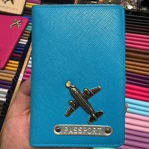 Könnte beinhalten: Ein türkisfarbener Reisepasshalter mit strukturierter Oberfläche. Er hat ein bronzefarbenes Flugzeugemblem und eine Metallplatte mit der Aufschrift "PASSPORT". Im Hintergrund sind weitere Reisepasshalter in verschiedenen Farben zu sehen.