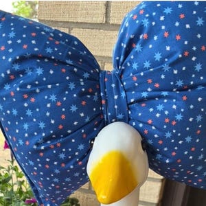 Puede incluir: Una cabeza de ganso decorativa con un gran lazo azul. El lazo tiene un estampado de estrellas blancas, azules y rojas. La cabeza del ganso es blanca con un pico amarillo. El lazo está atado alrededor del cuello del ganso.