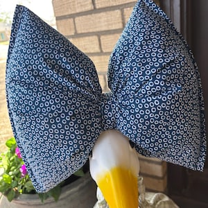 May include: A decorative goose figurine with a large navy blue bow featuring a white circle pattern. The goose has a white head, a bright yellow beak, and is wearing a floral patterned dress. The background includes a brick wall and a potted plant.