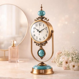 May include: A decorative gold-colored desk clock with a round face and black hands. The clock features a teal-colored base and accents, with a matching teal sphere at the top and a dangling teal sphere below the clock face. The clock is an elegant home decor item.