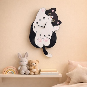 May include: A whimsical wall clock shaped like a black and white cat with a round clock face. The clock features black hands and pink paw prints. A shelf below holds a stuffed bunny, teddy bear, and a rainbow-shaped wooden toy.