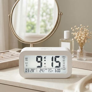 May include: A digital alarm clock displays the time as 9:15 AM, with the date and temperature also visible. The clock is white with a rectangular shape and is placed on a light-colored surface. A mirror and decorative items are in the background.