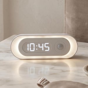 May include: A white and gray digital alarm clock with a rounded rectangular shape. The clock displays the time 10:45 in white digits. The clock has a motion sensor and a glowing light around the perimeter.
