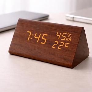 May include: A wooden, triangular alarm clock displaying the time 7:45, 45% humidity, and 22 degrees Celsius. The clock has a dark brown finish and a digital display with orange numerals. The clock is on a light-colored surface.