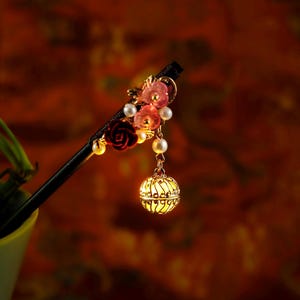 Puede incluir: Un palillo de pelo negro adornado con flores rojas y rosas, perlas y un orbe de filigrana dorada. El arreglo floral incluye una rosa roja y flores rosas. El orbe cuelga de una cadena, añadiendo elegancia a este accesorio para el cabello.