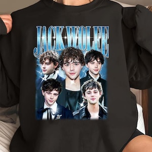 Puede incluir: Una sudadera negra de cuello redondo con un estampado gráfico de cinco retratos de la misma persona de pelo oscuro. Sobre los retratos, el texto JACK WOLFE está impreso en una fuente azul de estilo metálico sobre un fondo de rayos azules.