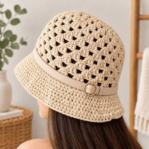 May include: A beige crochet bucket hat featuring an openwork pattern throughout the crown and brim. The hat is accented with a smooth, matching beige faux leather strap that wraps around the base, secured by a small, circular metal buckle.