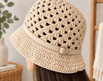 Op de afbeelding: Een beige gehaakte bucket hat met een opengewerkt patroon over de gehele kroon en rand. De hoed is geaccentueerd met een gladde, bijpassende beige kunstlederen band die rond de basis loopt, vastgezet met een kleine, ronde metalen gesp.