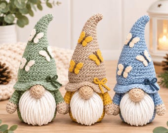 Op de afbeelding: Drie handgemaakte gehaakte kabouters op een rij. De linker kabouter is saliegroen met witte vlinders, de middelste is beige met gele vlinders en de rechter is lichtblauw met witte vlinders. Elke kabouter heeft een witte baard van garen.