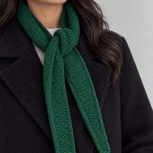 May include: A dark green knitted scarf with a textured garter stitch pattern, tied in a simple knot around the neck. The scarf is worn over a white ribbed sweater and a black wool coat, featuring clean edges and a consistent deep forest green color.
