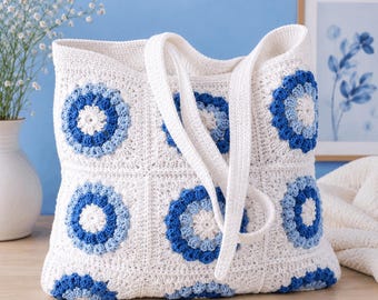 Op de afbeelding: Een witte gehaakte tote bag met een patroon van vierkante motieven. Elk vierkant bevat een cirkelvormig ontwerp met concentrische ringen in tinten wit, lichtblauw en donkerblauw. De tas heeft twee witte gehaakte schouderbanden aan de bovenzijde.