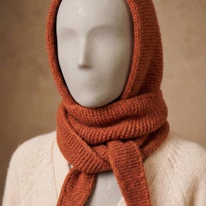 Op de afbeelding: Een roestkleurige gebreide bivakmuts met een geïntegreerde sjaal die om de nek wikkelt. Het accessoire heeft een consistente ribbelsteektextuur over het geheel, met twee lange, taps toelopende uiteinden die aan de voorzijde naar beneden hangen.