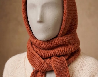 Op de afbeelding: Een roestkleurige gebreide bivakmuts met een geïntegreerde sjaal die om de nek wikkelt. Het accessoire heeft een consistente ribbelsteektextuur over het geheel, met twee lange, taps toelopende uiteinden die aan de voorzijde naar beneden hangen.