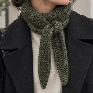 May include: An olive green knitted scarf with a textured garter stitch pattern, tied in a simple knot at the neck. The scarf is worn over a cream-colored sweater and underneath a black coat, featuring tapered ends that hang down the chest.