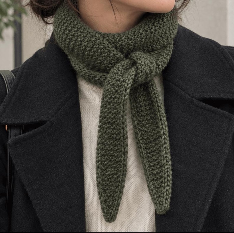 May include: An olive green knitted scarf with a textured garter stitch pattern, tied in a simple knot at the neck. The scarf is worn over a cream-colored sweater and underneath a black coat, featuring tapered ends that hang down the chest.