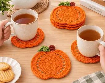 Op de afbeelding: Een set oranje pompoenvormige gehaakte onderzetters met groene blaadjes en een bruin steeltje. De onderzetters zijn rond met een decoratief opengewerkt patroon. Twee onderzetters liggen onder keramische mokken met thee, terwijl een stapel van drie andere onderzetters in de buurt op een houten oppervlak ligt.