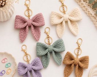 Op de afbeelding: Vijf gehaakte strik-sleutelhangers in mauve, crème, saliegroen, lavendel en bruin. Elke strik is bevestigd aan een goudkleurige metalen karabijnhaak en een ronde sleutelring. De gestructureerde garen strikken hebben een centrale knoop.