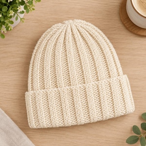 Könnte beinhalten: Eine cremefarbene Strickmütze mit vertikaler Rippenstruktur über das gesamte Modell und einem breiten, umgeschlagenen Bündchen an der Basis. Die Mütze ist aus dickem Garn mit einem gleichmäßigen Strickmuster und einer abgerundeten Krone gefertigt.