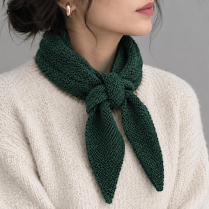 May include: A dark forest green knitted scarf tied in a simple knot around the neck. The scarf features a textured, garter stitch pattern with pointed ends. It is worn over a cream-colored, long-sleeved, chunky knit sweater with a ribbed texture.