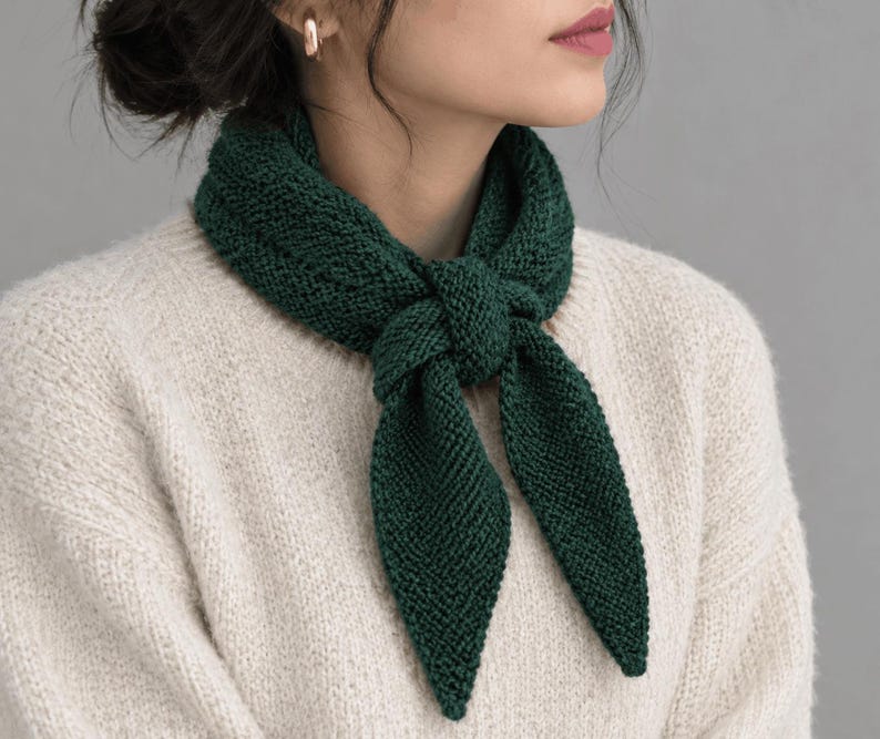 May include: A dark forest green knitted scarf tied in a simple knot around the neck. The scarf features a textured, garter stitch pattern with pointed ends. It is worn over a cream-colored, long-sleeved, chunky knit sweater with a ribbed texture.