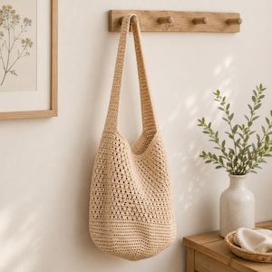 Puede incluir: Un bolso hobo de ganchillo en color beige que presenta un patrón texturizado de punto abierto en todo el cuerpo y una base sólida y de tejido apretado. El bolso incluye una sola correa ancha para el hombro integrada en la estructura.