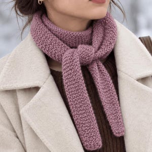 May include: A mauve knitted scarf with a textured garter stitch pattern, tied in a simple knot around the neck. The scarf is worn over a dark brown ribbed sweater and a cream-colored wool coat with wide lapels and a brown shoulder strap.